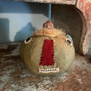 May include: A round, olive green fabric pumpkin decoration featuring a dark red fabric nose patch, a cream-coloured stitched mouth, and two black button eyes. The top is adorned with a textured brown stem and a tuft of light brown raffia fibres.