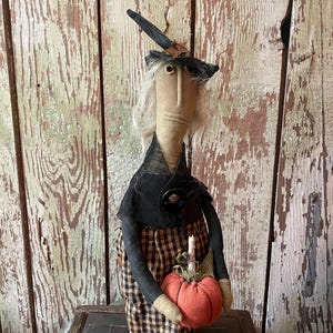 May include: A fabric doll shaped like a witch with a tall pointed black hat, grey yarn hair, and a tan face. The doll wears a black shawl with a dark button and a black and tan checkered skirt, while holding a small orange fabric pumpkin.