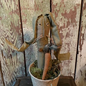 May include: A handcrafted, rustic bunny figure with long, drooping ears and a carrot. The bunny is made of olive green fabric and has button eyes and a twig-like moustache. It sits in a textured pot filled with moss.