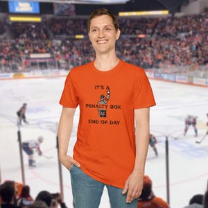 May include: Orange t-shirt with a black and white illustration of a hockey referee signaling a penalty. The text on the shirt reads "It's a Penalty Box Kind of Day."