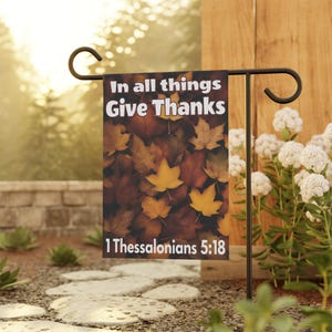 Może przedstawiać: Prostokątna flaga ogrodowa z napisem "In all things Give Thanks" i "1 Thessalonians 5:18". Flaga ma tło z jesiennych liści w odcieniach brązu i żółci. Jest prezentowana na czarnym metalowym stojaku.