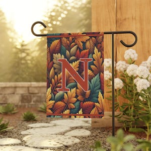 Könnte beinhalten: Eine Gartenflagge mit einem farbenfrohen Herbstblattmuster und dem Buchstaben "N" in Rostfarbe. Die Flagge wird auf einem schwarzen Metallständer in einem Garten mit einem Steinweg und Grün in der Umgebung präsentiert.