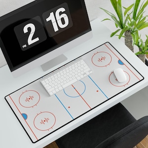 May include: A desk mat with a hockey rink design, featuring red and blue lines and circles on a white background. A white keyboard and mouse are on the mat, with a computer monitor showing the time. A plant is visible.