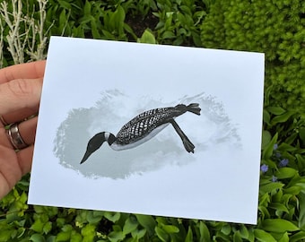 May include: A greeting card with a monochrome illustration of a loon in flight. The bird is shown with detailed plumage and a long, pointed beak. The background is a soft, grey wash, and the card is held against a backdrop of green foliage.