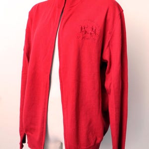 May include: A vibrant red zip-up sweater with a high collar. The sweater features a small embroidered emblem on the chest. The zipper is black. The sweater is displayed on a mannequin.