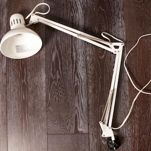 May include: White adjustable desk lamp with a round shade and articulated arm. The lamp has a white cord and a black base. The lamp is on a dark wood surface.