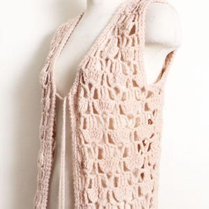 May include: A light pink, open-front crochet vest. The vest is sleeveless and has a long, flowing design. The crochet pattern features an open, lacy design. The vest is displayed on a mannequin. The vest has a tie closure with tassels.