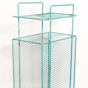 May include: A tall, rectangular, mint green metal mesh storage stand featuring a wire grid construction, two open shelves, and four thin legs with rounded feet. The structure is composed of thin metal rods and a fine wire mesh throughout.
