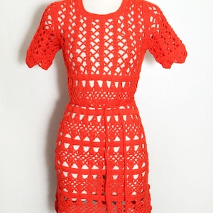 May include: A vibrant red crochet dress displayed on a mannequin. The dress features a round neckline, short sleeves, and a fitted waist. The skirt has a scalloped hemline. The open weave design reveals the mannequin beneath.