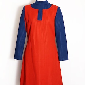 May include: A sleeveless red dress with a subtle vertical ribbed texture, featuring a square neckline and a hem that falls above the knee. The dress is layered over a long-sleeved, ribbed navy blue turtleneck top, creating a color-blocked look.