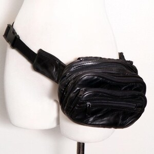 May include: A black leather fanny pack featuring multiple zippered compartments and a black adjustable nylon strap with a plastic side-release buckle. The waist bag has a rounded, multi-pocket design with visible stitching and metal zipper pulls.