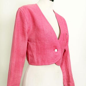 May include: A cropped, pink blazer with long sleeves and a single-button closure. The blazer features a V-neck and a small pocket on the front. The garment is displayed on a mannequin.