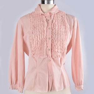 May include: A pale pink, long-sleeved blouse with a ruffled collar and front panel. The blouse has a button-up closure and a fitted silhouette.