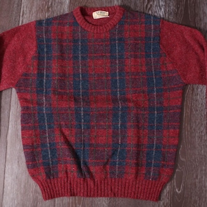 May include: A red sweater with a plaid pattern in shades of red, blue, and green. The sweater has long sleeves and a ribbed hem and cuffs. The neckline is a crew neck.