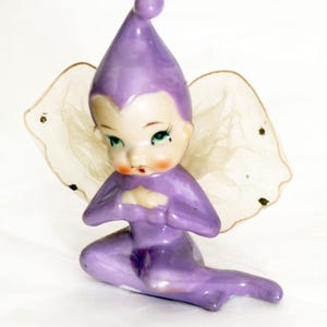 May include: A ceramic figurine of a seated elf wearing a purple pointed hat and matching purple outfit. The elf has green eyes, rosy cheeks, and translucent, off-white mesh wings with small dark accents attached to the back of the shoulders.
