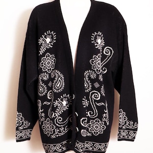 May include: Black cardigan sweater with white paisley and floral patterns. The open-front sweater features a V-neck and long sleeves. The cuffs and hem have a decorative border. The sweater is displayed on a mannequin.