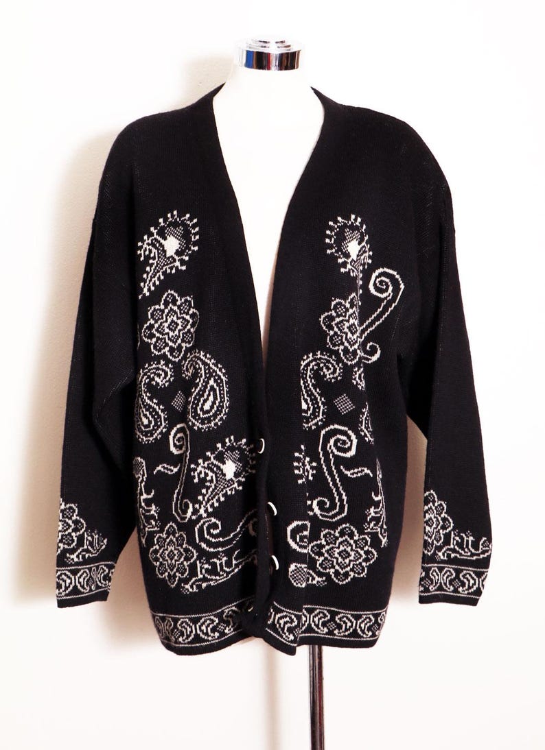 May include: Black cardigan sweater with white paisley and floral patterns. The open-front sweater features a V-neck and long sleeves. The cuffs and hem have a decorative border. The sweater is displayed on a mannequin.