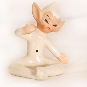 May include: A ceramic elf figurine with a glossy off-white finish, pointed ears, and a matching cap. The figure is seated with legs crossed, one arm raised, and features painted black eyes, rosy cheeks, and a small circular hole on the torso.