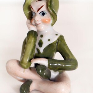 May include: A small ceramic figurine of a seated elf wearing a green hat, a green long-sleeved top with white accents and green spots, and green shoes. The elf has a pale complexion, rosy cheeks, and is resting one hand against its chin.