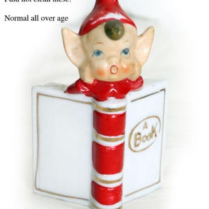 May include: A ceramic figurine of an elf with a red hat and collar, positioned behind a white book with a red and white striped spine. The book features gold trim and the text A Book in a gold oval. The elf has blue eyes and a small green dot.