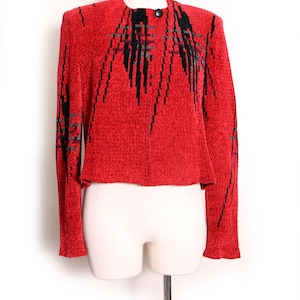 May include: A red knit jacket with black and gray geometric designs. The jacket has a round neckline, long sleeves, and a single black button closure. The garment is displayed on a mannequin.