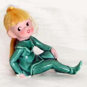 May include: A ceramic figurine of a seated elf with a glossy dark green outfit, pointed shoes, and a collar. The figure has a pale face with rosy cheeks, a small red mouth, and synthetic yellow hair tied in a high ponytail at the top of the head.
