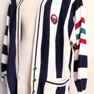 May include: A navy blue and white striped cardigan. The V-neck sweater has a button closure and two front pockets. The sleeve has red and green stripes. An embroidered emblem is on the chest.