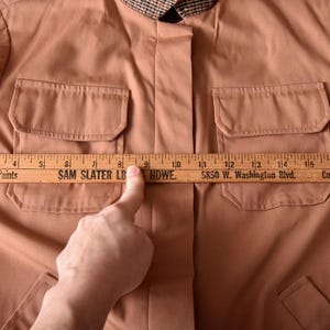May include: A brown jacket with two large pockets on the front. A wooden ruler is placed across the front of the jacket, measuring 10 inches across the chest. The ruler has the text "Benjamin Moore Paints", "SAM SLATER LB HDWE", "5850 W. Washington Blvd.", "Culver City", and "WE 3-5724" printed on it.