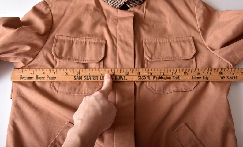 May include: A brown jacket with two large pockets on the front. A wooden ruler is placed across the front of the jacket, measuring 10 inches across the chest. The ruler has the text "Benjamin Moore Paints", "SAM SLATER LB HDWE", "5850 W. Washington Blvd.", "Culver City", and "WE 3-5724" printed on it.