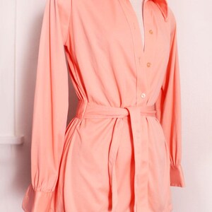 May include: A peach-colored vintage shirt dress with long sleeves, a collared neckline, and a matching belt. The dress features button closures down the front and is displayed on a mannequin. The sleeves have a cuff detail.