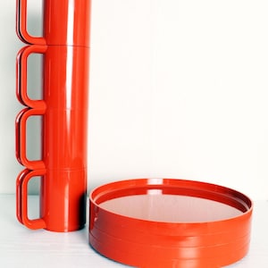 Puede incluir: Una pila de cuatro tazas de color rojo brillante con asas grandes y curvas, junto a una bandeja redonda roja a juego. Las tazas son cilíndricas y apiladas verticalmente. La bandeja tiene un perfil bajo con un borde ligeramente elevado. Los artículos son de material brillante.