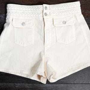 Puede incluir: Pantalones cortos color crema de cintura alta con cinturilla trenzada y dos bolsillos delanteros. Los pantalones cortos tienen dos cierres de botón y están hechos de una tela resistente. Los pantalones cortos son de color neutro y se pueden combinar con muchas blusas.
