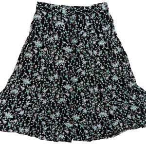 Puede incluir: Falda negra con estampado floral y botones en la parte delantera. La falda presenta un diseño escalonado y un patrón de pequeñas flores azules y rosas. La falda está hecha de una tela ligera.
