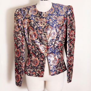 May include: A long-sleeved jacket featuring a vibrant floral brocade pattern in shades of blue, red, and gold. The garment has structured shoulders, a round neckline, and a front closure secured by four circular gold-toned buttons with dark centers.