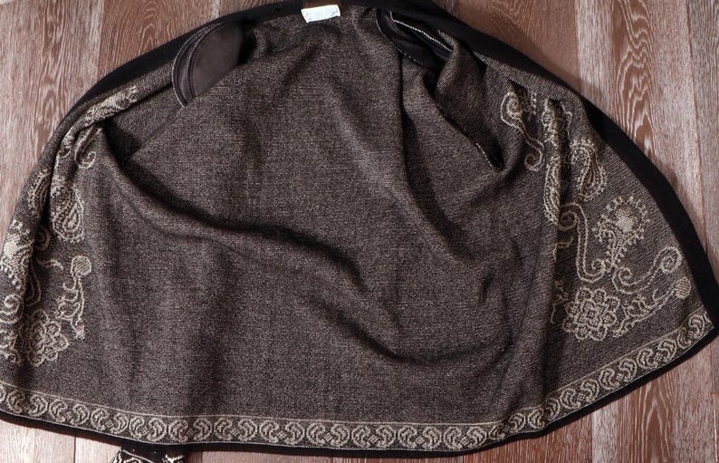 May include: A black jacket with a dark gray interior. The interior features an ornate, cream-colored paisley design along the edges. The jacket is open, revealing the lining and the intricate pattern.