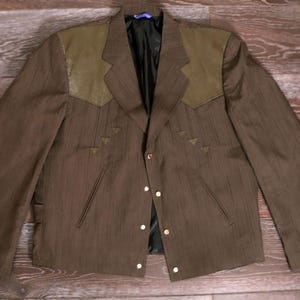 May include: Brown blazer with olive green shoulder accents and gold buttons. The blazer has a textured fabric and a classic design, suitable for various occasions. Features a notched lapel and decorative stitching.