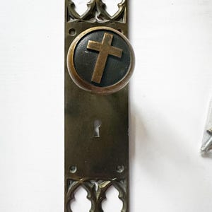 May include: A brass door knob and rectangular backplate set featuring a dark circular inlay on the knob with a centered brass cross. The backplate has decorative pointed arches at the top and bottom and a central keyhole opening.