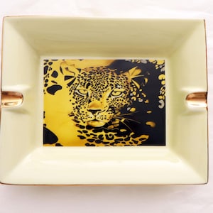 Op de afbeelding: Rechthoekige asbak in crèmekleur met gouden rand. Het midden toont een gezicht van een luipaard in geel en zwart. De asbak heeft twee ingebouwde steunen voor sigaretten. Een decoratief item voor in huis.