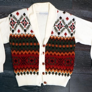 Könnte beinhalten: Cremefarbene Strickjacke mit geometrischem Muster in Rot, Grün, Schwarz und Orange. Die Jacke hat einen Schalkragen, lange Ärmel und Knopfverschlüsse. Das Muster wiederholt sich auf der Brust und am unteren Teil der Jacken.