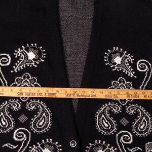 May include: Black cardigan sweater with white paisley design. The sweater measures 23.5 inches across. The open front cardigan has a V-neck and two small buttons. The sweater is made of a soft material.