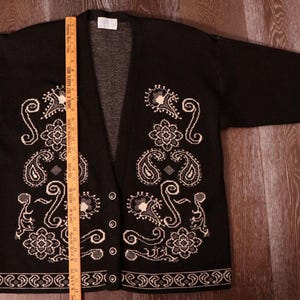 May include: Black cardigan sweater with white paisley and floral embroidery. The open-front sweater has long sleeves and decorative trim on the cuffs and hem. The label reads "28" tall".