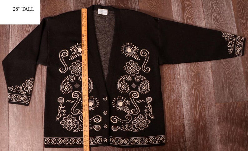 May include: Black cardigan sweater with white paisley and floral embroidery. The open-front sweater has long sleeves and decorative trim on the cuffs and hem. The label reads "28" tall".