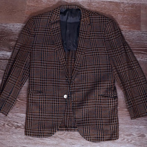 Könnte beinhalten: Ein braun-schwarzer Blazer mit Karomuster, einem Ein-Knopf-Verschluss und zwei Vordertaschen. Die Jacke hat ein klassisches Design und ist aus einem gewebten Stoff gefertigt. Der Blazer liegt flach auf einer Holzoberfläche.