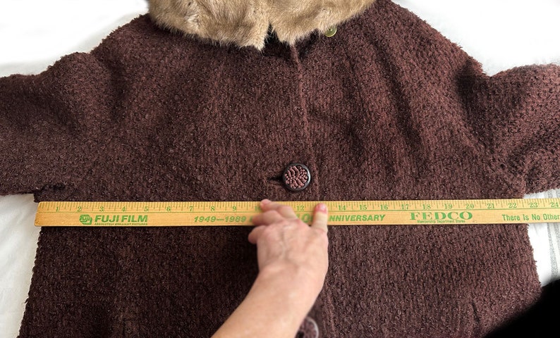 May include: A brown fuzzy coat with a fur collar. The coat is being measured with a wooden ruler that reads "FUJI FILM 1949-1989 50th ANNIVERSARY FEDCO There Is No Other"