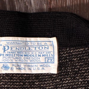 May include: Close-up of a black and white knit fabric with a blue and white label. The label reads "WARRANTED TO BE A PENDLETON" and includes text about the fabric's composition and origin. The label also indicates the size is 2X.