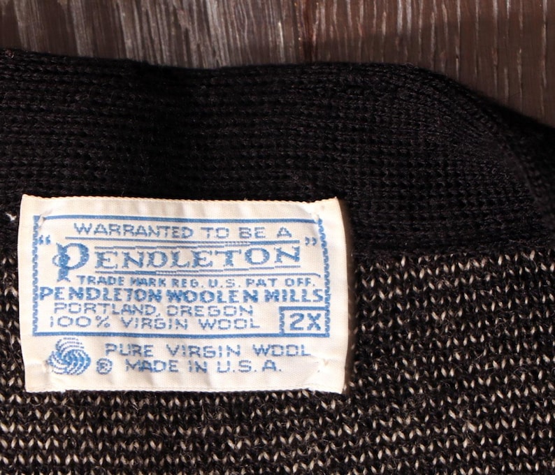 May include: Close-up of a black and white knit fabric with a blue and white label. The label reads "WARRANTED TO BE A PENDLETON" and includes text about the fabric's composition and origin. The label also indicates the size is 2X.
