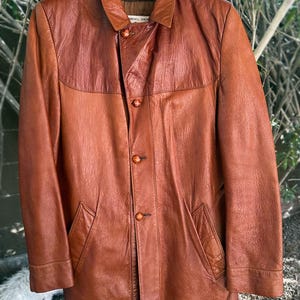 May include: A brown leather jacket with a collared neckline and button-down closure. The jacket features two front pockets and a two-tone design, with a darker brown yoke and collar. The jacket is hanging on a wooden hanger.