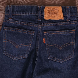 Puede incluir: Primer plano de la parte trasera de unos vaqueros de mezclilla azul oscuro con costuras naranjas. Cuenta con dos bolsillos traseros, una pequeña etiqueta naranja y una etiqueta marrón de Levi Strauss & Co.