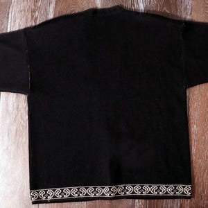 May include: Black long-sleeve shirt with white floral embroidery on the cuffs and hem. The shirt is laid flat on a wooden surface, showcasing the design details. The sleeves are extended, displaying the full length of the garment.