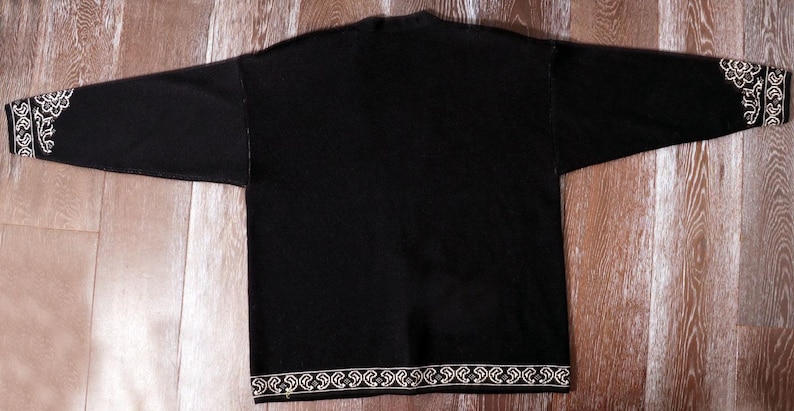 May include: Black long-sleeve shirt with white floral embroidery on the cuffs and hem. The shirt is laid flat on a wooden surface, showcasing the design details. The sleeves are extended, displaying the full length of the garment.